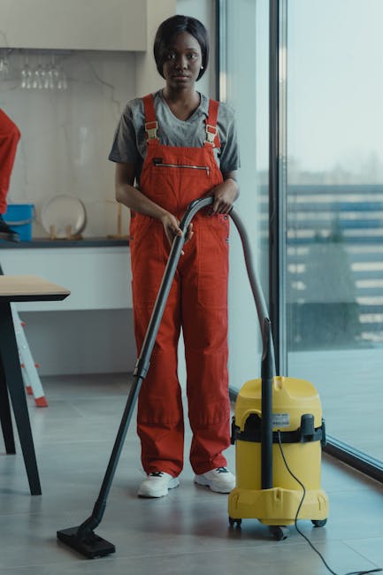 A woman dressed in casual grey top and bright red overalls stands in a modern kitchen area, operating a yellow industrial vacuum cleaner connected to a long black hose, which she uses for surface clea