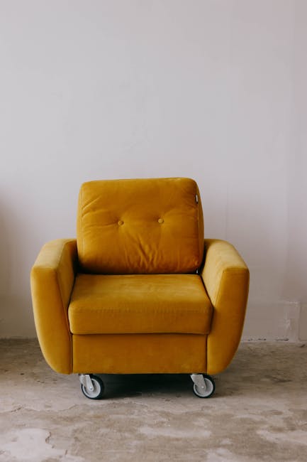 A mustard yellow upholstered armchair with a mid-century modern design, featuring a tufted backrest and wide armrests, positioned on a concrete floor in front of a plain white wall. The chair has smal